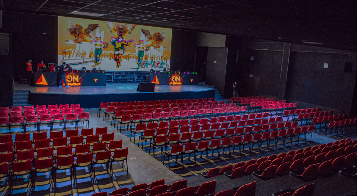 Auditório com cadeiras vermelhas e palco iluminado, apresentando um cenário colorido e equipamentos de som. O auditório utiliza barreiras acústicas e materiais absorventes para melhorar a qualidade sonora e reduzir a poluição sonora.