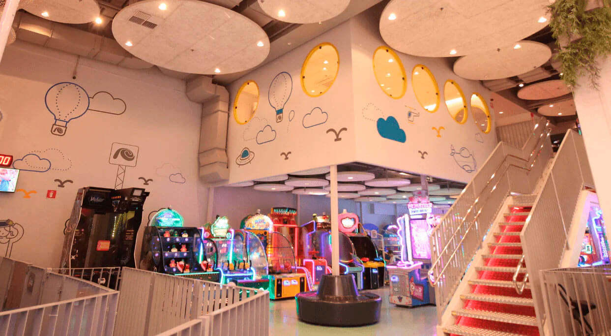 Área de recreação interna com vários jogos eletrônicos e máquinas de arcade coloridas. O teto é decorado com grandes nuvens acústicas, que integram o isolamento acústico com a arquitetura do espaço.