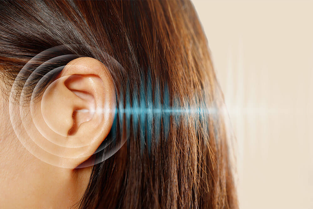 Visão lateral da cabeça de uma mulher, focando em sua orelha. Ondas sonoras ilustradas em azul e branco estão direcionadas para o ouvido, simbolizando audição ou possíveis problemas auditivos.