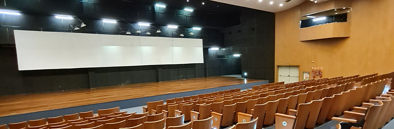 Interior de um auditório moderno. O palco é grande e está localizado à esquerda, com um fundo preto e paredes de madeira clara. Ele conta com revestimentos acústicos na parede e teto.