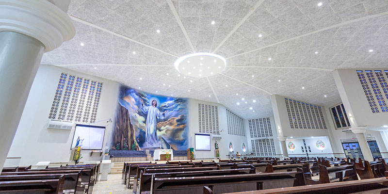 Interior de uma igreja com um amplo altar central. Acima do altar, há uma grande pintura de Jesus Cristo em uma paisagem celestial. O teto da igreja é alto e feito forro acústico com várias luzes embutidas, formando um padrão radial ao redor de um grande lustre central.