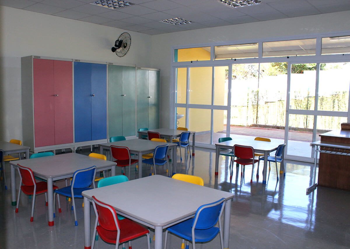 Sala de aula moderna com mesas e cadeiras coloridas, armários de cores suaves (rosa, azul e verde), e amplas janelas de vidro que permitem a entrada de luz natural. O piso é liso e brilhante, e há um ventilador de parede instalado no canto superior.