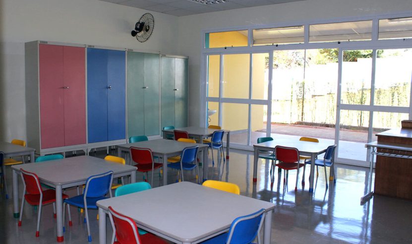 Sala de aula moderna com mesas e cadeiras coloridas, armários de cores suaves (rosa, azul e verde), e amplas janelas de vidro que permitem a entrada de luz natural. O piso é liso e brilhante, e há um ventilador de parede instalado no canto superior.