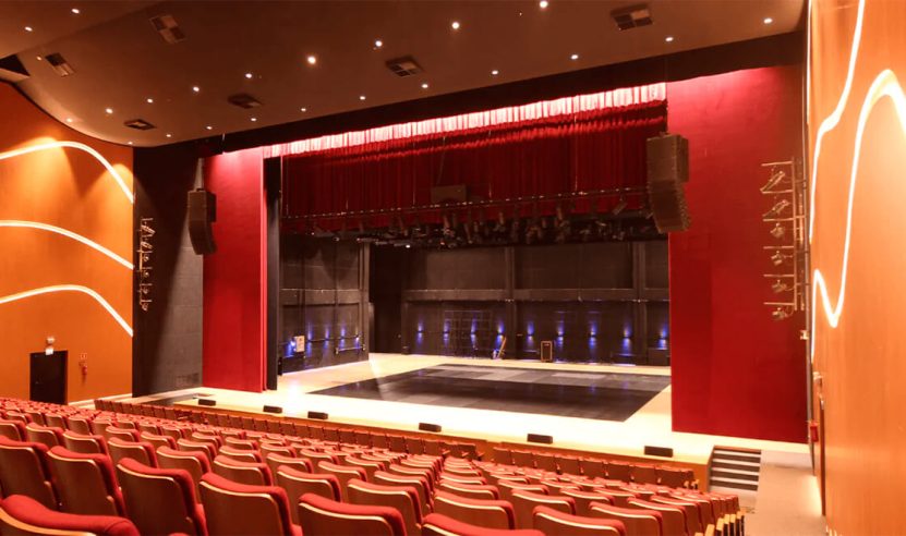 Teatro iluminado com cadeiras vermelhas e um palco equipado com luzes de espetáculo. As paredes e o teto apresentam painéis acústicos para melhorar a qualidade sonora do ambiente.