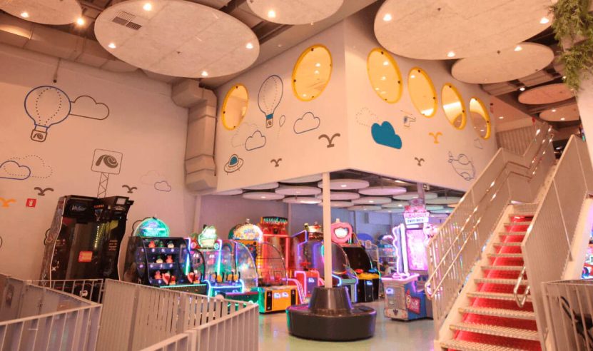 Área de recreação interna com vários jogos eletrônicos e máquinas de arcade coloridas. O teto é decorado com grandes nuvens acústicas, que integram o isolamento acústico com a arquitetura do espaço.