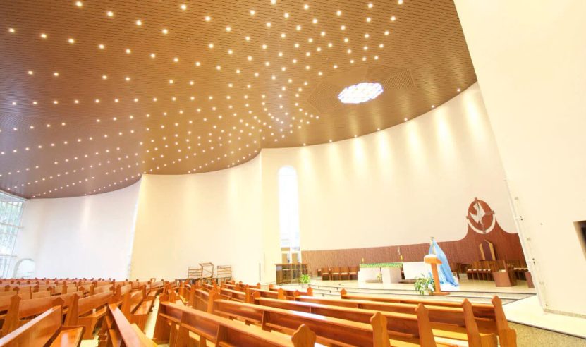 Igreja com teto alto e revestido de painéis acústicos de madeira, com iluminação embutida. Interior moderno com bancos de madeira e altar ao fundo, criando um ambiente acolhedor e acusticamente tratado.
