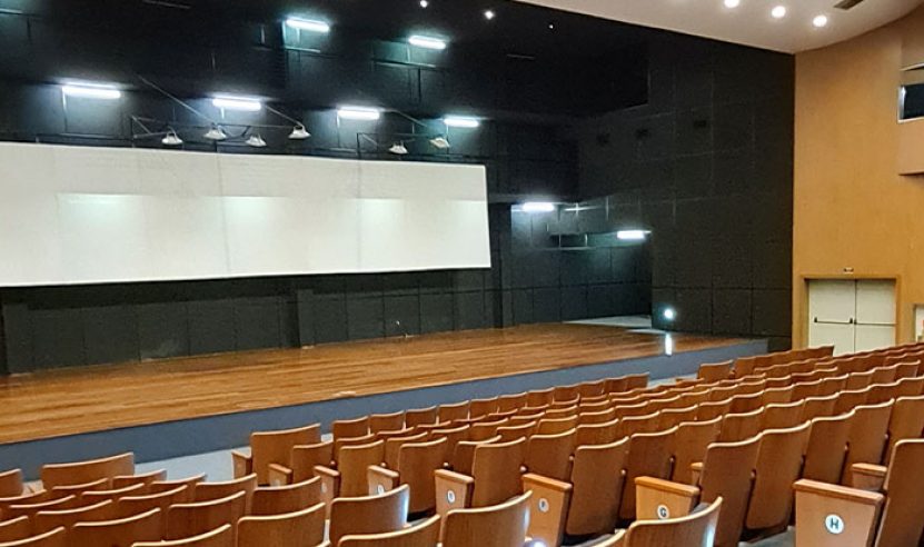 Interior de um auditório moderno. O palco é grande e está localizado à esquerda, com um fundo preto e paredes de madeira clara. Ele conta com revestimentos acústicos na parede e teto.