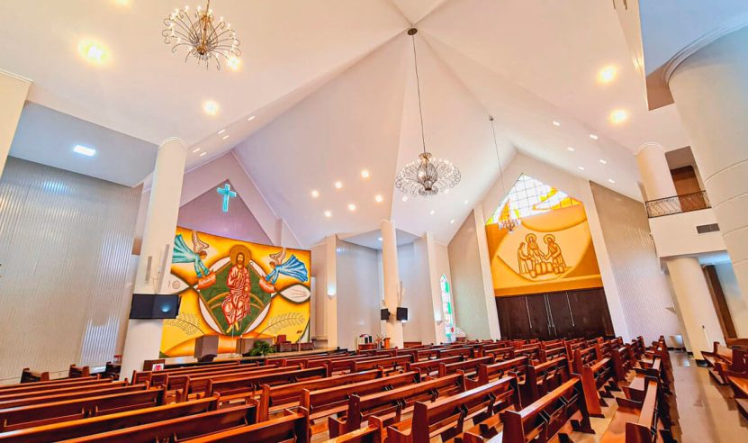 Interior de uma igreja moderna com um teto alto e inclinado. No fundo, há um grande mural colorido que representa figuras religiosas, incluindo Jesus no centro. À direita, há outra representação de figuras religiosas em um painel amarelo. O espaço é iluminado por várias luzes de teto e candelabros decorativos. Os bancos de madeira estão alinhados em fileiras, voltados para o altar. O ambiente é claro e bem iluminado, com uma atmosfera acolhedora e reverente.