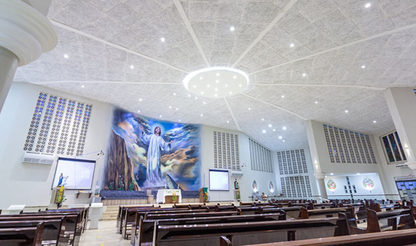 Interior de uma igreja com um amplo altar central. Acima do altar, há uma grande pintura de Jesus Cristo em uma paisagem celestial. O teto da igreja é alto e feito forro acústico com várias luzes embutidas, formando um padrão radial ao redor de um grande lustre central.