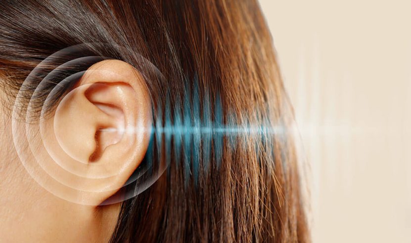 Visão lateral da cabeça de uma mulher, focando em sua orelha. Ondas sonoras ilustradas em azul e branco estão direcionadas para o ouvido, simbolizando audição ou possíveis problemas auditivos.